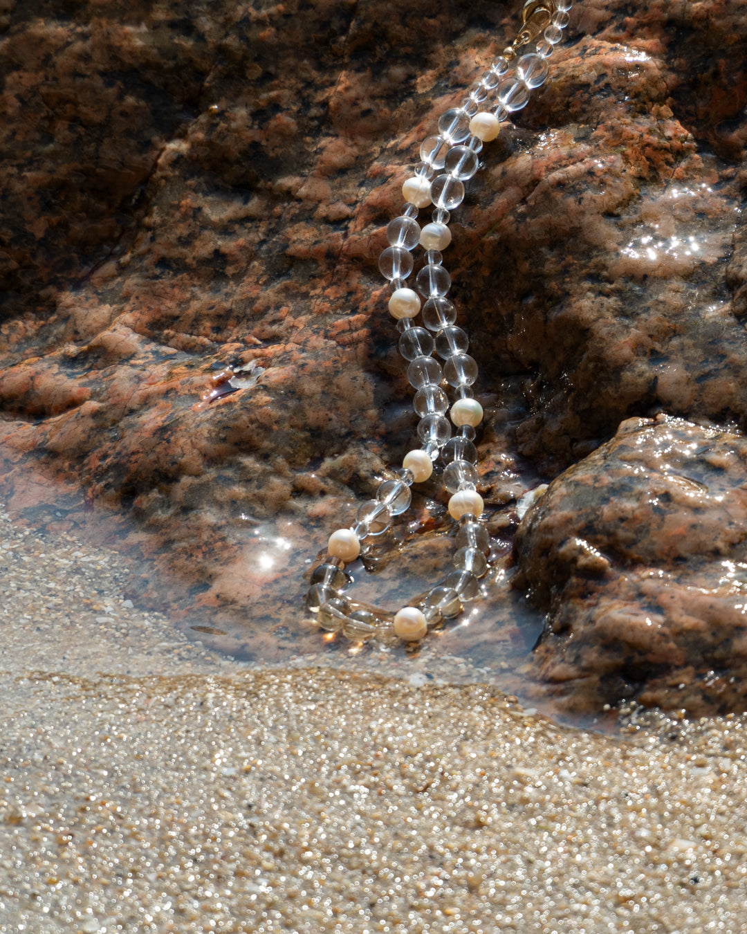 Elegant, Clear Quartz and Freshwater Pearl Eterna Nova necklace on rocks by water, showcasing delicate jewelry with natural beauty.