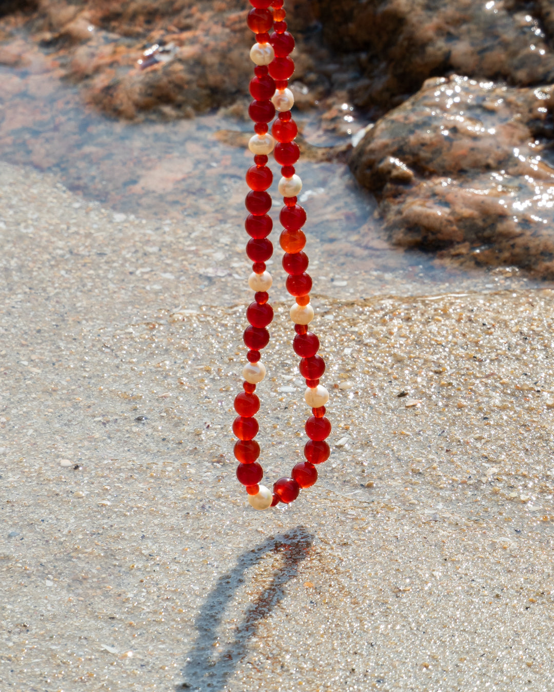 An Eterna Nova necklace made from vibrant red agate gemstones and freshwater pearls, elegantly resting on a rocky shore.