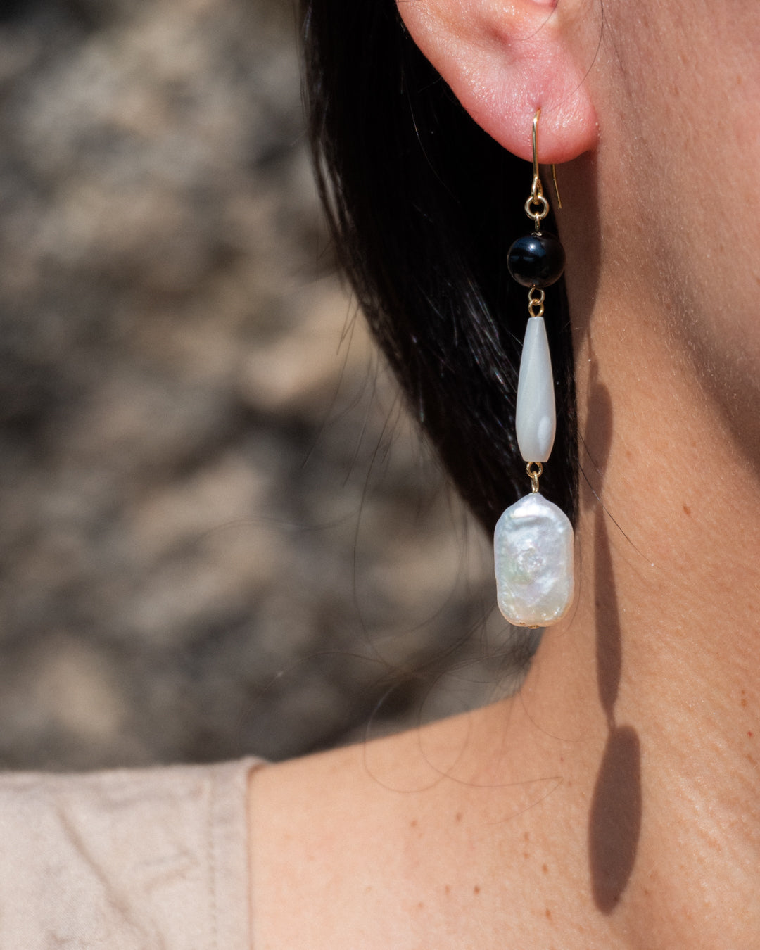 Furin earrings of freshwater baroque pearl, mother of pearl (MOP), and onyx, worn in front of a rocky background.