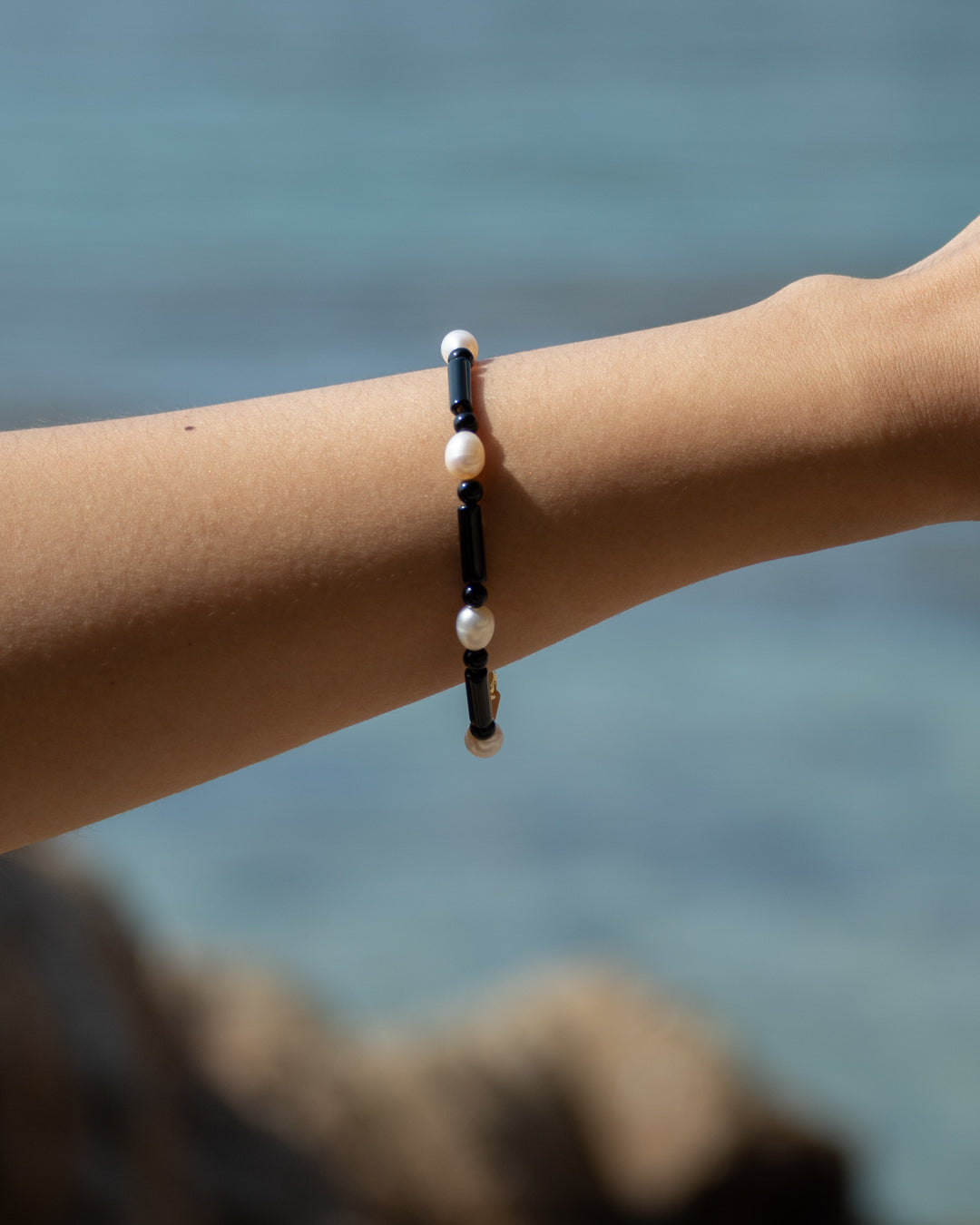 A person wearing a Starry Bracelet made with onyx and freshwater pearls, set against an ocean backdrop.