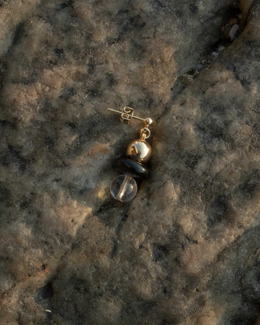 14k gold vermeil Stella Earrings featuring onyx button and clear quartz beads, set against a rocky background. The earring is designed to showcase the sparkle of the gemstones.