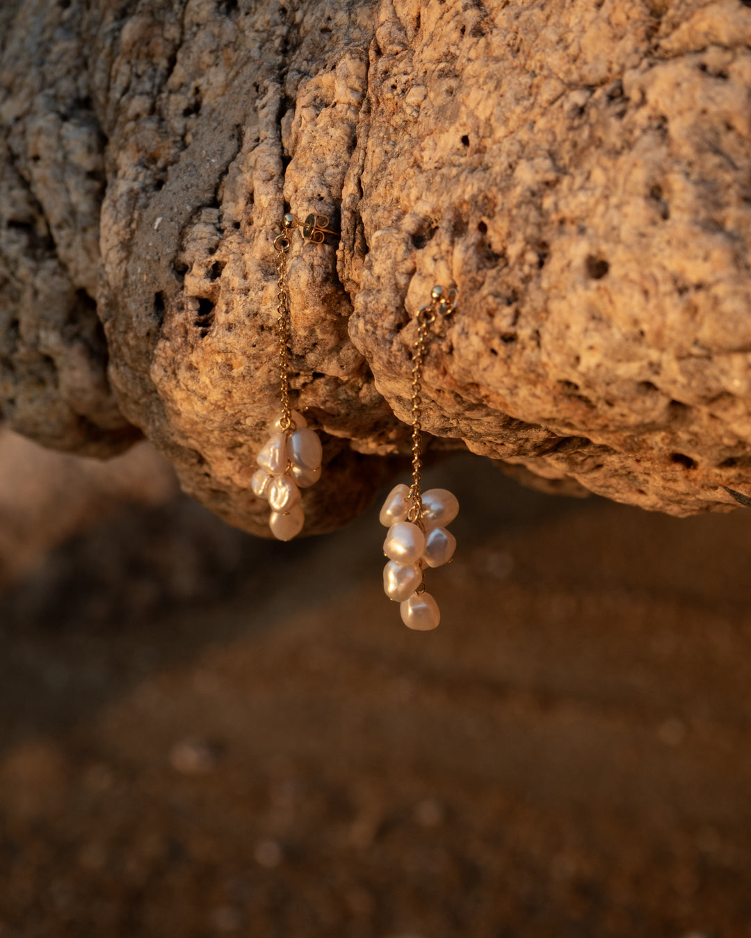 Floral Bloom Earrings - Freshwater Keshi Pearl Earring with gold vermeil chain. The jewelry features a delicate arrangement of pearls and is suspended from the sturdy gold chain, hanging against the backdrop of a natural stone wall. Perfect for fashion enthusiasts, this piece combines elegance with a touch of nature's charm.