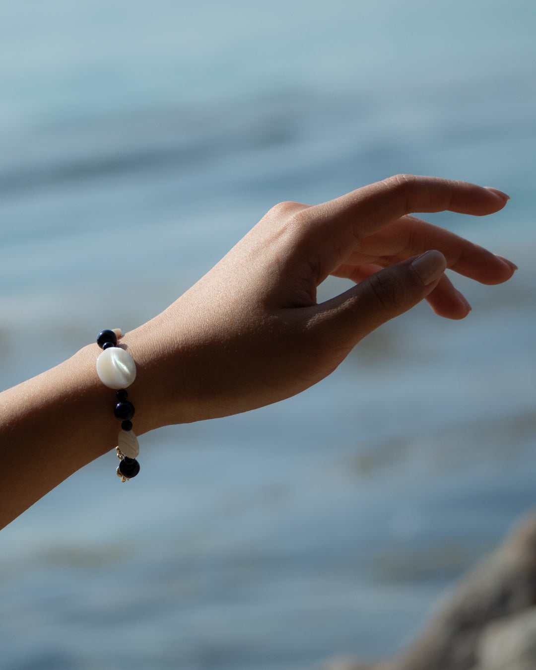 A person's hand showcasing a Sonata Moonlit Onyx Bracelet, set against a backdrop of the ocean, highlighting the onyx and mother-of-pearl stones.