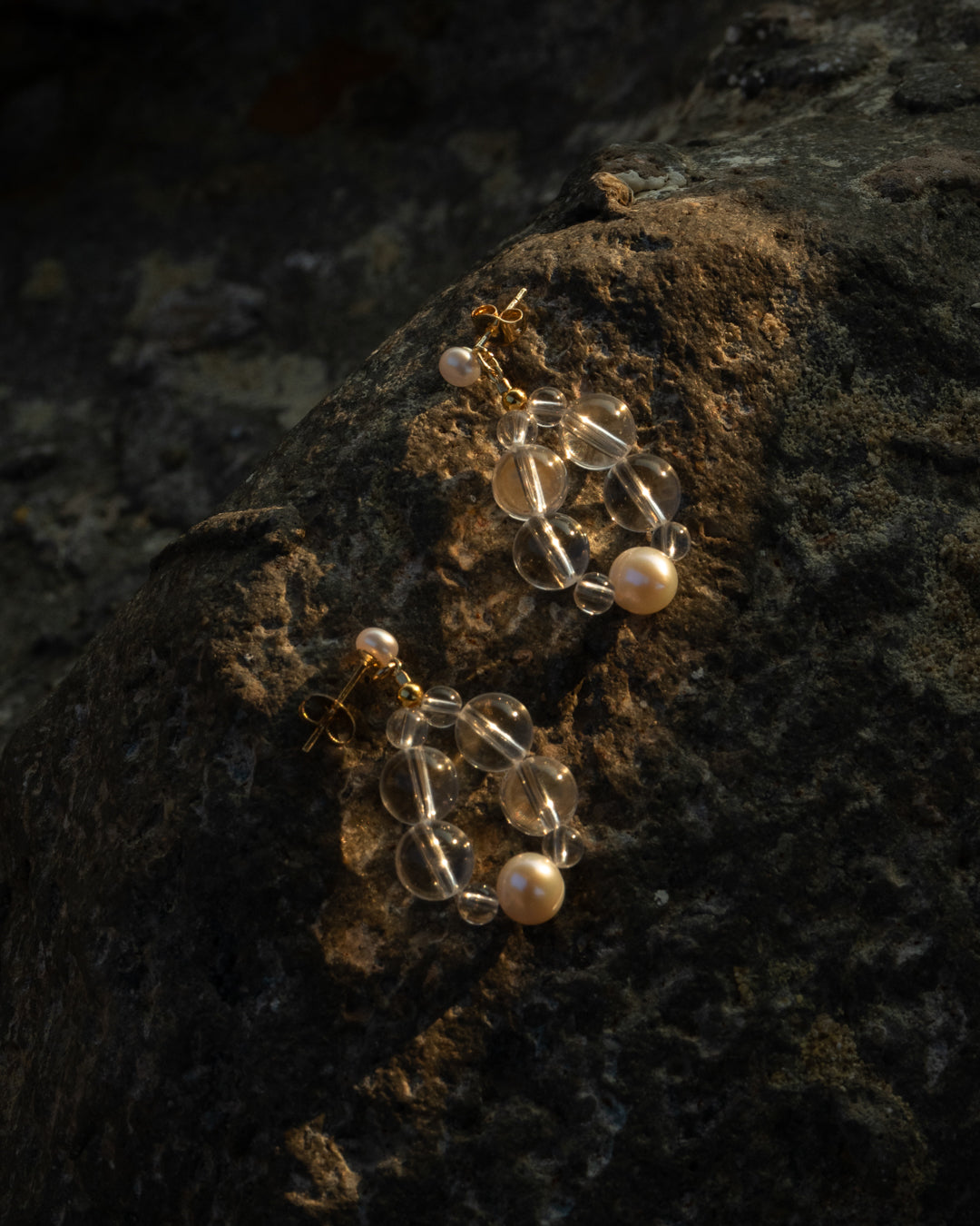 Freshwater pearl and clear quartz Eterna Nova earrings: Small, sparkling jewelry featuring natural gemstones and freshwater pearls set against a rocky background highlighted by the last ray of sunlight, showcasing their elegance.