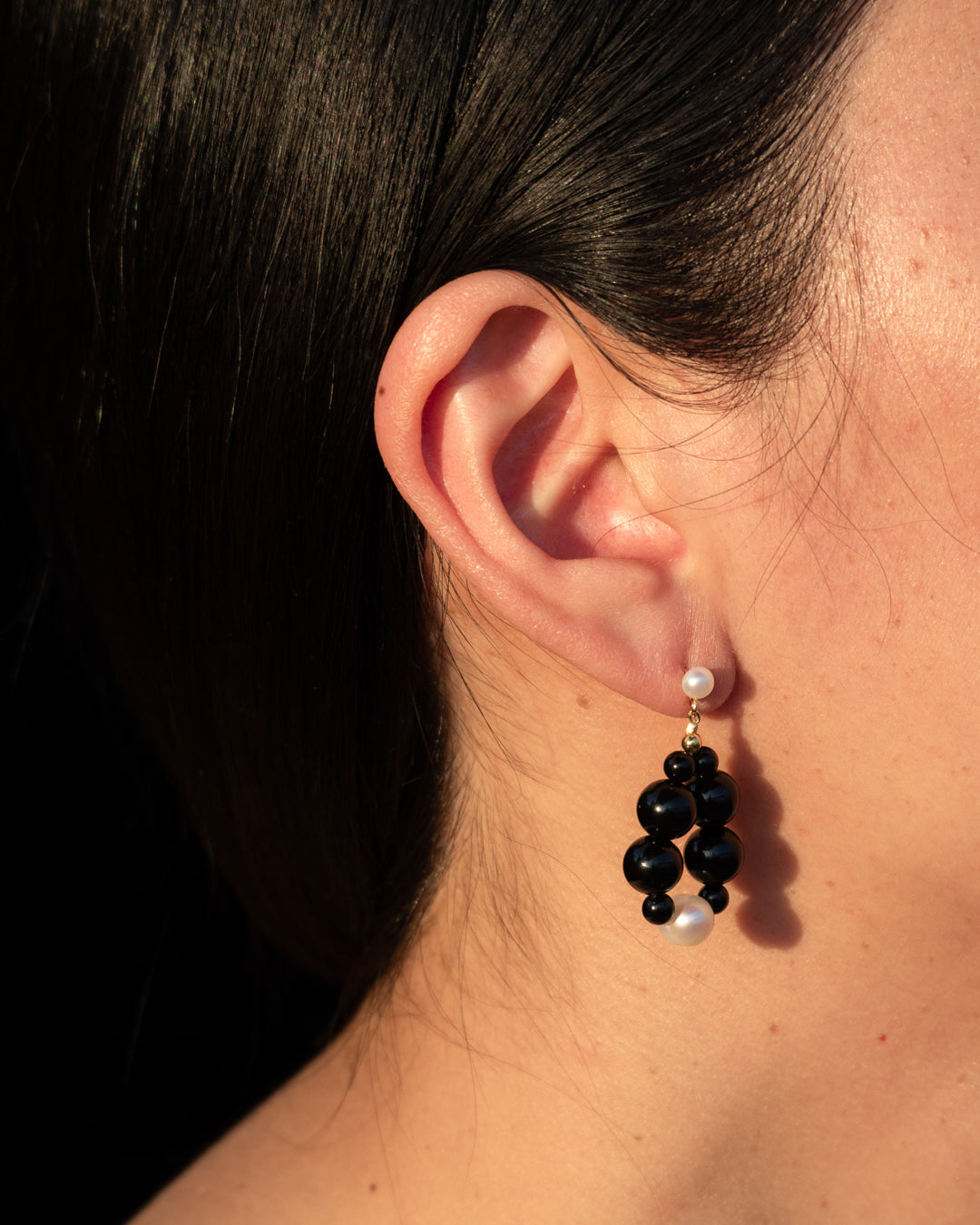 Elegant Eterna Nova Earrings onyx (small) featuring freshwater pearls and onyx, set against a black background, displayed by a woman with her hair pulled back. 