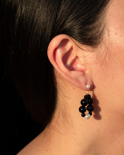 Elegant Eterna Nova Earrings onyx (small) featuring freshwater pearls and onyx, set against a black background, displayed by a woman with her hair pulled back. 