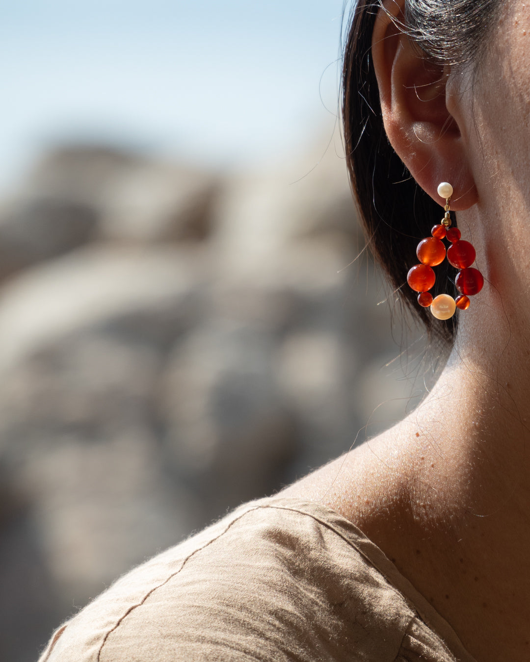 Elegant Eterna Nova Red Agate(Small) Earrings, showcasing their freshwater pearl accents, on a Pearsons ear with the rocky beach/ocean background, capturing the jewelry's intricate details and craftsmanship.