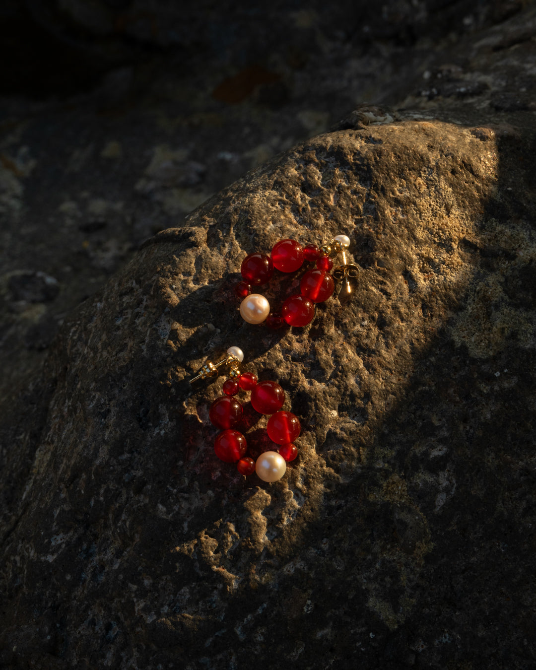Eterna Nova(small) earrings features red agate and freshwater pearls, on a rocky surface graced by the last ray of the sunlight of that day, showcasing their unique design against a rock backdrop. 