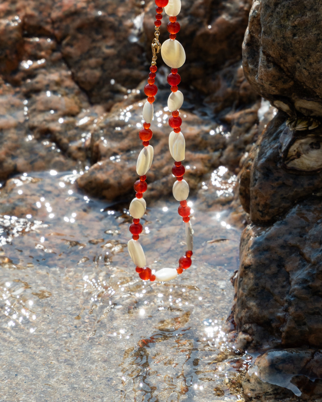 Vivid red agate beads against soft mother-of-pearl pieces, hanging above ocean water against a rocky beach background, evoking a sense of natural beauty and serenity.