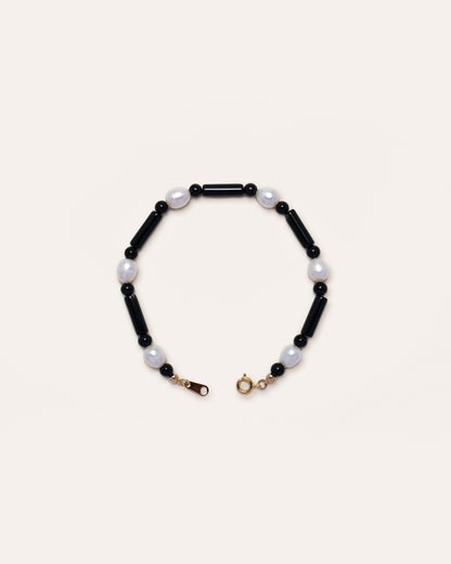 Starry Bracelet: A delicate black bracelet with onyx and freshwater pearl beads, featuring a gold clasp, presented against a minimalist grey background. 